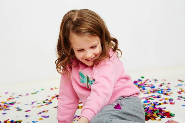 Joyeux belle petite fille appréciant surprise confettis colorés tombant, posant sur le mur de studio blanc. Jolie fille fête son anniversaire, s'amuser — Photo