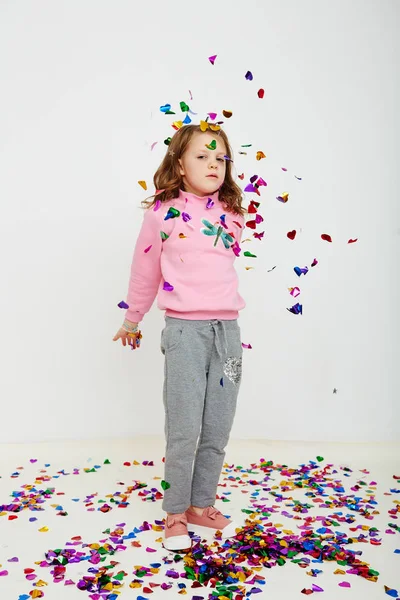 Happy beautiful little girl enjoying colorful confetti surprise falling down, posing on white studio wall. Pretty girl celebrating her birthday party, having fun — Stock Photo, Image