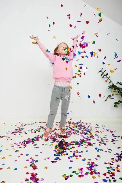 Feliz hermosa niña disfrutando de colorida sorpresa confeti cayendo, posando en la pared del estudio blanco. Chica bonita celebrando su fiesta de cumpleaños, divirtiéndose — Foto de Stock