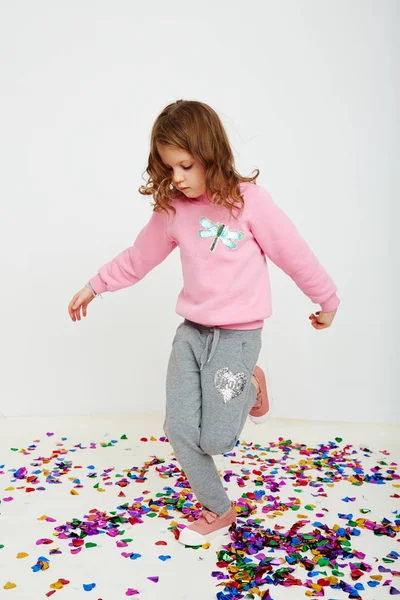 Happy beautiful little girl enjoying colorful confetti surprise falling down, posing on white studio wall. Pretty girl celebrating her birthday party, having fun — Stock Photo, Image