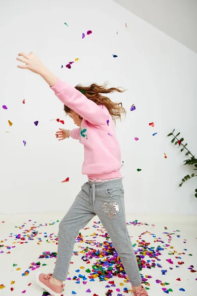 Happy beautiful little girl enjoying colorful confetti surprise falling down, posing on white studio wall. Pretty girl celebrating her birthday party, having fun