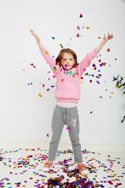 Menina bonita feliz desfrutando de confete colorido surpresa caindo, posando na parede do estúdio branco. Menina bonita comemorando sua festa de aniversário, se divertindo — Fotografia de Stock