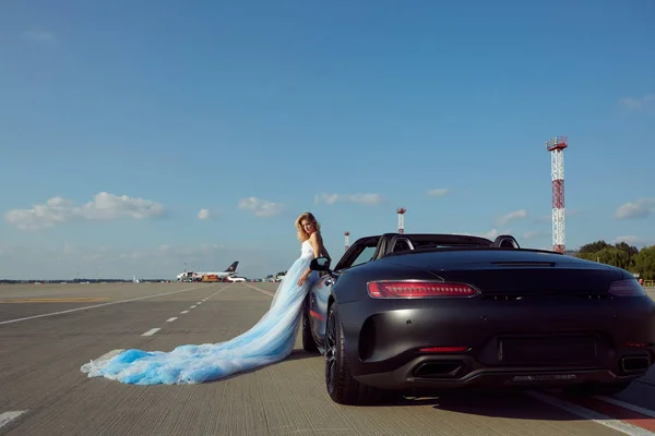 The elegant blonde beautiful woman posing near luxury vehicle on planes background. Girl wearing blue dress. No retouch. — ストック写真