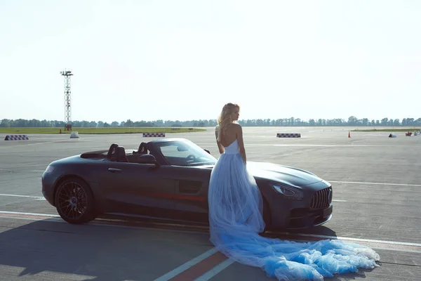 The elegant blonde beautiful woman posing near luxury vehicle on planes background. Girl wearing blue dress. No retouch. — ストック写真
