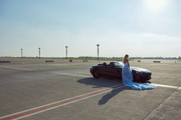 De elegante blonde mooie vrouw poseren in de buurt van luxe voertuig op vliegtuigen achtergrond. Een meisje in een blauwe jurk. Geen retoucheren. — Stockfoto