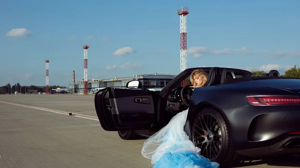 Элегантная блондинка красивая женщина позирует возле роскошного автомобиля на фоне аэропорта. Девушка в синем платье. Без повторного прикосновения — стоковое фото