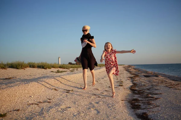 Madre sorridente e bellissima figlia si divertono sulla spiaggia. Ritratto di donna felice che dà un passaggio a cavalluccio a simpatica bambina con spazio copia. Ritratto di bambina e sua madre durante l'estate — Foto Stock