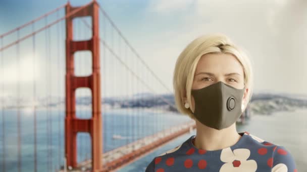 Portræt af en smuk blond ung kvinde iført beskyttende medicinsk ansigtsmaske og stående foran Golden Gate Bridge. Online rejse koncept – Stock-video