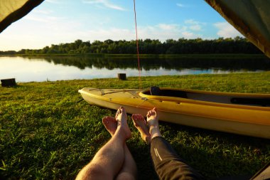 Çimenli bir çayırda ve nehir kıyısında çadırdan çıkan iki çift bacak..