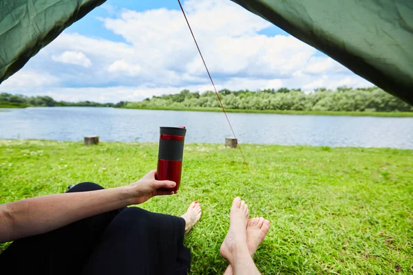 Dois pares de pernas sobressaem da tenda em um prado de clareira gramado e perto do rio. — Fotografia de Stock