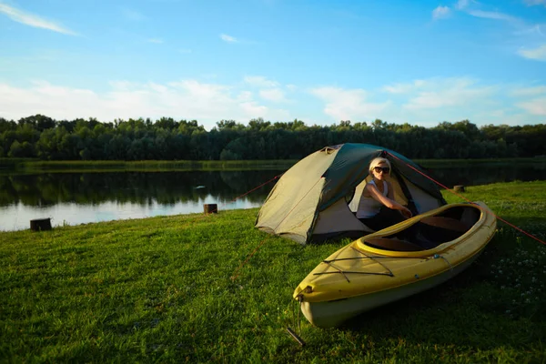 Perjalanan, kayak dan hiking konsep. Potret wanita cantik duduk di dekat tenda hijau dengan kayak di dekat sungai. — Stok Foto