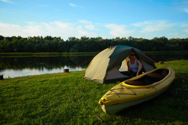 Travel, kayaking and hiking concept. Portrait of young beautiful woman sitting near green tent with kayak near river. — Stock Photo, Image