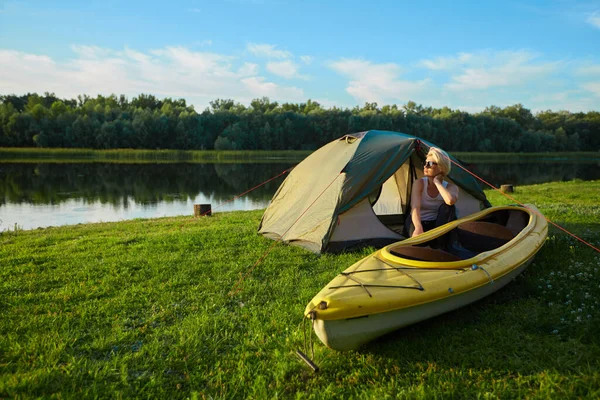 Travel, kayaking and hiking concept. Portrait of young beautiful woman sitting near green tent with kayak near river. — Stock Photo, Image