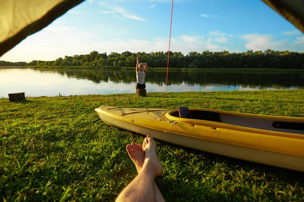 Young woman wakes up and stretches near the tent set on a coast of a river — Stock Photo, Image