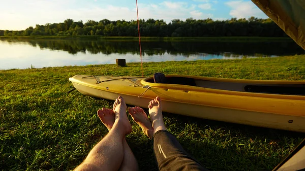 Két pár láb áll ki a sátorból egy füves réten és a folyó közelében.. — Stock Fotó