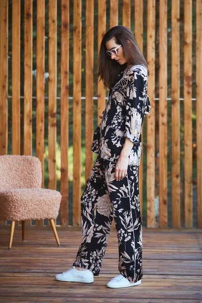 Young stylish sexy woman in pajamas standing on terrace in tropical hotel, palm trees background, long black hair, sunglasses, earrings, sunglasses. — Stock Photo, Image