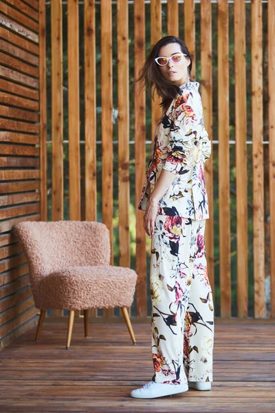 Giovane donna sexy elegante in pigiama in piedi sulla terrazza in hotel tropicale, sfondo di palme, lunghi capelli neri, occhiali da sole, orecchini, occhiali da sole. — Foto Stock