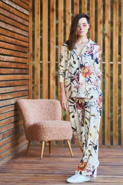 Jonge stijlvolle sexy vrouw in pyjama staande op terras in tropisch hotel, palmbomen achtergrond, lang zwart haar, zonnebril, oorbellen, zonnebril. — Stockfoto