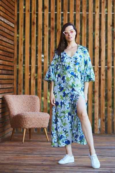 Ung snygg sexig kvinna i sommar klänning stående på trä terrass i tropiskt hotell, palmer bakgrund, långt svart hår, solglasögon, örhängen, solglasögon. — Stockfoto