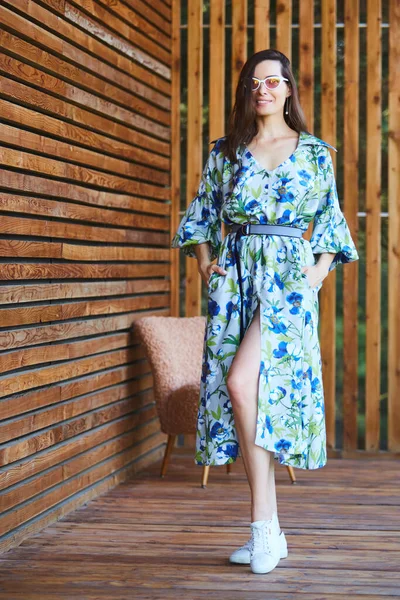 Junge stilvolle sexy Frau im Sommerkleid steht auf einer Holzterrasse im tropischen Hotel, Palmen Hintergrund, lange schwarze Haare, Sonnenbrille, Ohrringe, Sonnenbrille. — Stockfoto