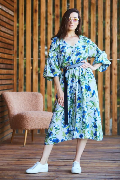 Mulher sexy elegante jovem em vestido de verão em pé no terraço de madeira em hotel tropical, palmeiras fundo, cabelo preto longo, óculos de sol, brincos, óculos de sol . — Fotografia de Stock