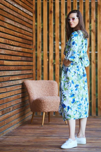 Mujer sexy con estilo joven en vestido de verano de pie en la terraza de madera en el hotel tropical, fondo de palmeras, pelo negro largo, gafas de sol, pendientes, gafas de sol . — Foto de Stock