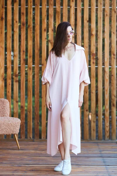 Mulher sexy elegante jovem em vestido rosa de verão em pé no terraço de madeira em hotel tropical, palmeiras fundo, cabelos longos e pretos, óculos de sol, brincos, óculos de sol . — Fotografia de Stock