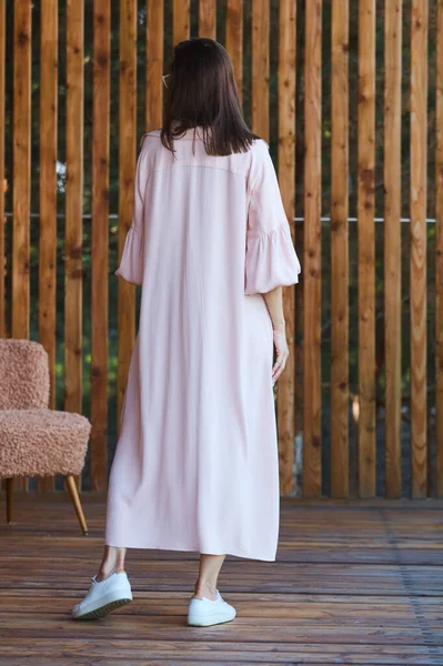 Mujer sexy con estilo joven en vestido rosa de verano de pie en la terraza de madera en el hotel tropical, fondo de palmeras, pelo largo y negro, gafas de sol, pendientes, gafas de sol . — Foto de Stock