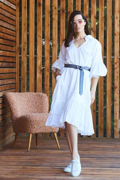 Mujer sexy con estilo joven en vestido blanco de verano de pie en la terraza de madera en el hotel tropical, fondo de palmeras, pelo largo y negro, gafas de sol, pendientes, gafas de sol — Foto de Stock