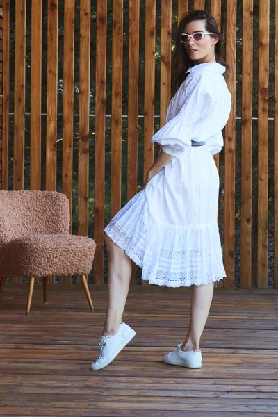 Mujer sexy con estilo joven en vestido blanco de verano de pie en la terraza de madera en el hotel tropical, fondo de palmeras, pelo largo y negro, gafas de sol, pendientes, gafas de sol —  Fotos de Stock