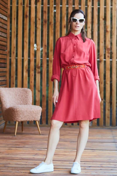 Jonge stijlvolle sexy vrouw in de zomer rode jurk staande op houten terras in tropisch hotel, palmbomen achtergrond, lang zwart haar, zonnebril, oorbellen, zonnebril — Stockfoto