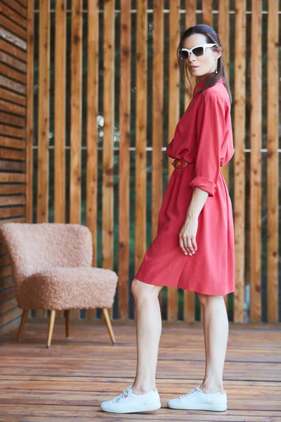 Mujer sexy con estilo joven en vestido rojo de verano de pie en la terraza de madera en el hotel tropical, fondo de palmeras, pelo largo y negro, gafas de sol, pendientes, gafas de sol —  Fotos de Stock