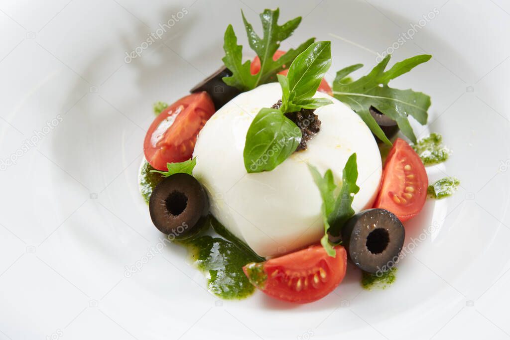 Traditional authentic soft and creamy fresh Italian burrata cheese shaped as white round ball over Italian tomatoes topped with rucola along side pesto reduction