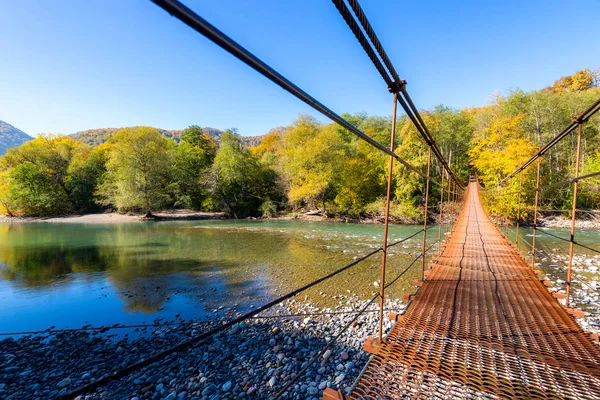 Narrow metal foot bridge across mountain river in autumn.
