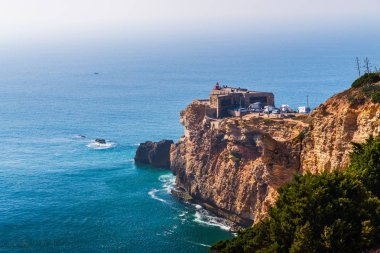 Portekiz 'in deniz kıyısındaki antik kale, Nazare. Kayalık kayalıklardaki deniz feneri. Atlantik Okyanusu kıyısı. Promontuarın yamaçları yeşil bitkilerle kaplıdır. Dalgalar kayalara çarpıyor..