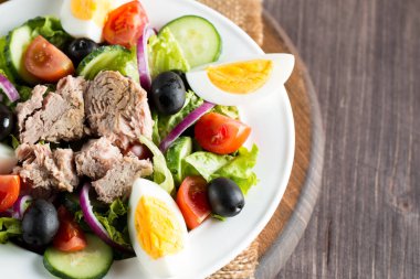 Domates, roka, ton balığı, yumurta, roka, kraker ve baharattan yapılmış taze balık salatası. Tahta arka planda beyaz bir kasede Sezar salatası.