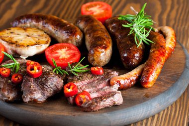 Karışık ızgara et tabağı yakın çekim fotoğraf. Sığır eti, domuz eti, kümes hayvanları, sosis, kızarmış sarımsak, biber biber, ahşap rustik arka plan üzerinde kırmızı domates.