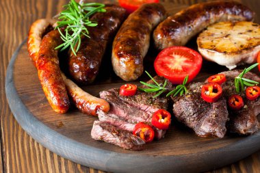 Karışık ızgara et tabağı yakın çekim fotoğraf. Sığır eti, domuz eti, kümes hayvanları, sosis, kızarmış sarımsak, biber biber, ahşap rustik arka plan üzerinde kırmızı domates.