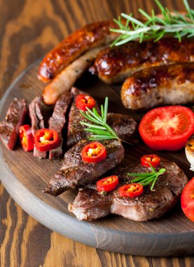 Karışık ızgara et tabağı yakın çekim fotoğraf. Sığır eti, domuz eti, kümes hayvanları, sosis, kızarmış sarımsak, biber biber, ahşap rustik arka plan üzerinde kırmızı domates.