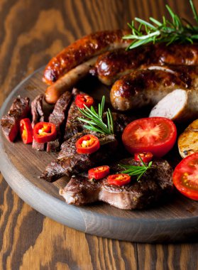 Karışık ızgara et tabağı yakın çekim fotoğraf. Sığır eti, domuz eti, kümes hayvanları, sosis, kızarmış sarımsak, biber biber, ahşap rustik arka plan üzerinde kırmızı domates.