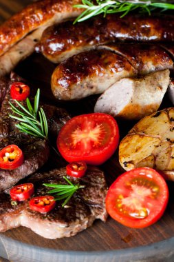 Karışık ızgara et tabağı yakın çekim fotoğraf. Sığır eti, domuz eti, kümes hayvanları, sosis, kızarmış sarımsak, biber biber, ahşap rustik arka plan üzerinde kırmızı domates.