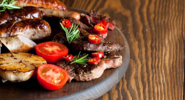 Karışık ızgara et tabağı yakın çekim fotoğraf. Sığır eti, domuz eti, kümes hayvanları, sosis, kızarmış sarımsak, biber biber, ahşap rustik arka plan üzerinde kırmızı domates.