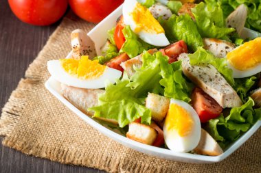 Taze salata domates, ruccola, tavuk göğsü, yumurta, roka, kraker ve baharat yapılmış. Ahşap arka plan üzerinde beyaz, şeffaf bir kasede Sezar salatası