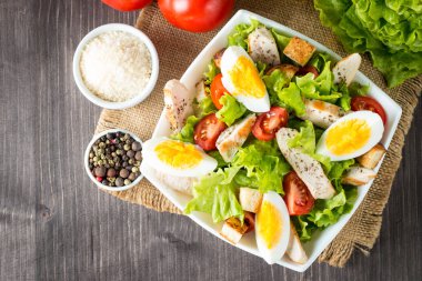 Taze salata domates, ruccola, tavuk göğsü, yumurta, roka, kraker ve baharat yapılmış. Ahşap arka plan üzerinde beyaz, şeffaf bir kasede Sezar salatası