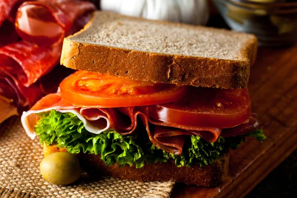 Close-up photo of a club sandwich. Sandwich with meat, prosciutto, salami, salad, vegetables, lettuce, tomato, onion and mustard on a fresh sliced rye bread on wooden background. Olives background.