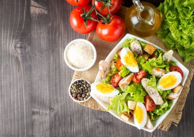 Taze salata domates, ruccola, tavuk göğsü, yumurta, roka, kraker ve baharat yapılmış. Ahşap arka plan üzerinde beyaz, şeffaf bir kasede Sezar salatası