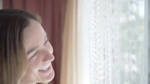 Menina feliz rindo enquanto olha para fora através da janela com cortinas, close-up — Vídeo de Stock