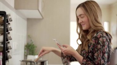 Gülümseyen genç kadın mutfakta fırında yemek pişirirken akıllı telefon kullanıyor.