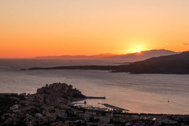Uzak dağların üzerinden yükselen güneş Korsika 'nın Balagne bölgesindeki Calvi kalesini ve körfezi aydınlatıyor.