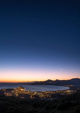 Hilal ay, güneş doğarken körfez üzerinde parlak bir gecede Korsika 'nın Balagne bölgesindeki Calvi kalesinde yamaç köylerinin ışıkları ve uzaktaki Kaptan Corse ile birlikte yükseliyor.